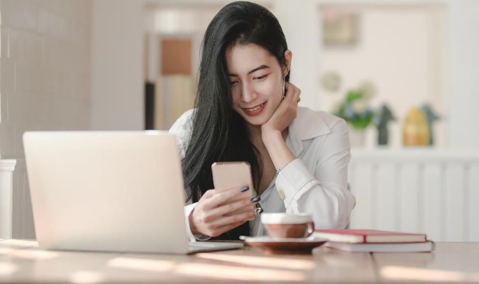 Female smiling whilst sitting in front of a laptop and texting on her phone. 