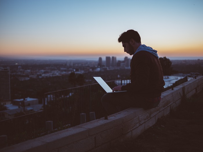 A young man works on a laptop while sitting on a city rooftop