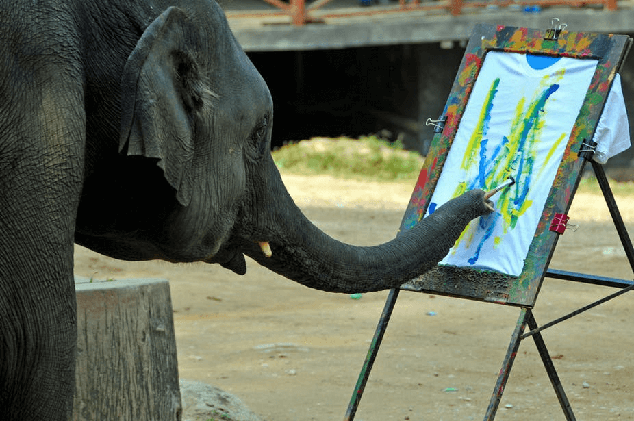An elephant with a paint brush in their trunk making marks on a piece of paper.